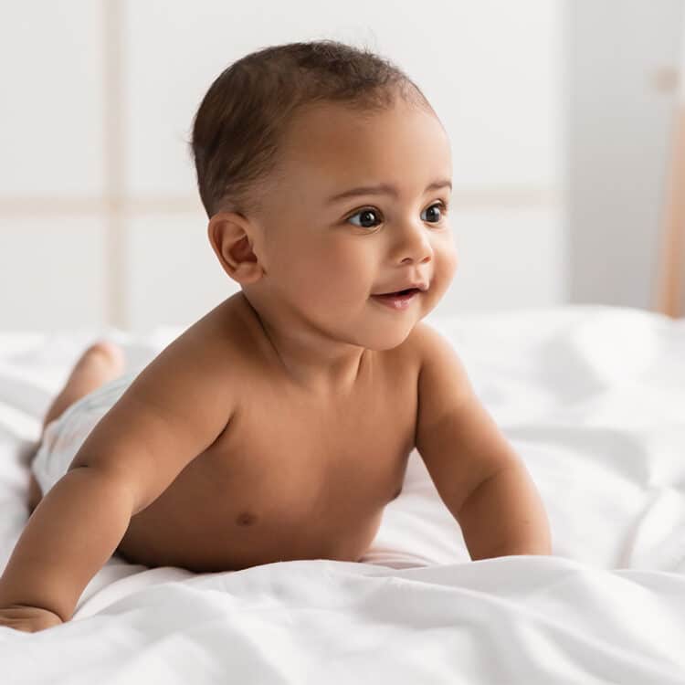 Imagem de um bebê de bruços em cima de um lençol branco, em cima da cama. Ele está apenas de fralda, tem a pele morena, olhos e cabelos escuros e está sorrindo.