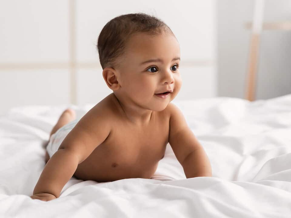 Imagem de um bebê de bruços em cima de um lençol branco, em cima da cama. Ele está apenas de fralda, tem a pele morena, olhos e cabelos escuros e está sorrindo.