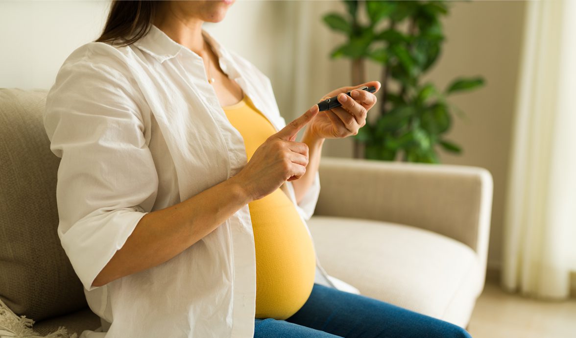 Imagem de uma gestante com diabetes gestacional, fazendo um teste de glicemia, por meio do furo no dedo indicador. A imagem está fechada no corpo da gestante, que está sentada em um sofá bege. Ela veste uma blusa amarela, com uma camisa branca por cima e calça jeans.