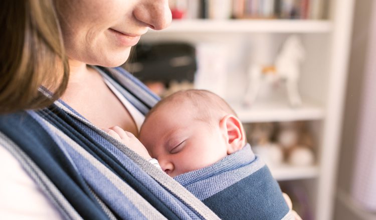 A imagem traz a uma mãe com seu bebê colado ao peito, dentro de um sling em tecido com listras largas nas cores azul e branco. Mãe e bebê tem pele clara e cabelos castanhos claro. O bebê está dormindo e a mãe olhando para ele e sorrindo levemente. O uso do sling é uma prática recomendada na exterogestação.