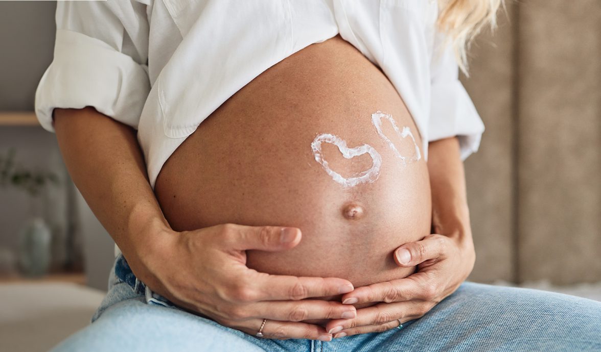 Imagem fechada na barriga de uma gestante. Ela tema pele levemente bronzeada e veste uma camisa branca e uma calça jeans. Está segurando a barriga com ambas as mãos e nela estão desenhados dois corações na cor branca, sinalizando que se trata de uma gravidez gemelar.