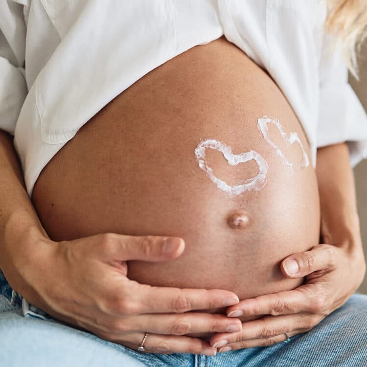 Imagem fechada na barriga de uma gestante. Ela tema pele levemente bronzeada e veste uma camisa branca e uma calça jeans. Está segurando a barriga com ambas as mãos e nela estão desenhados dois corações na cor branca, sinalizando que se trata de uma gravidez gemelar.