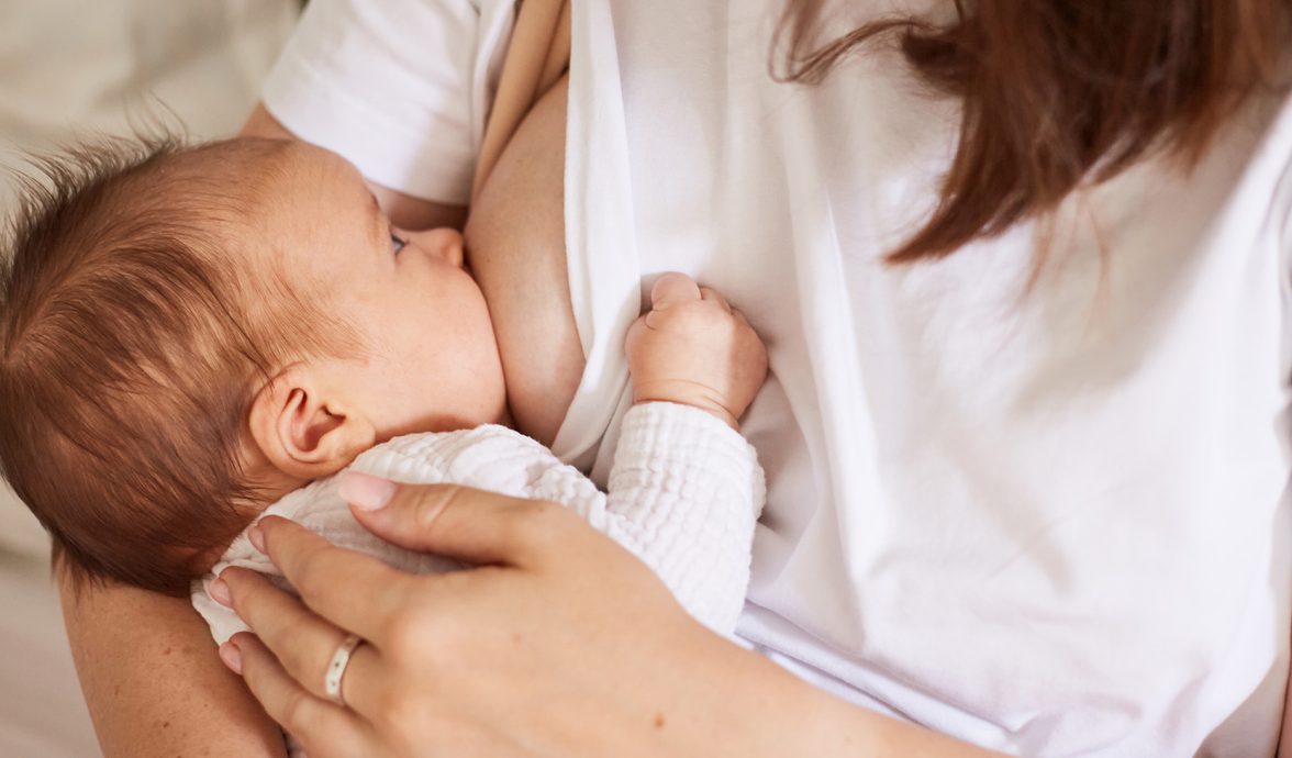 Imagem focada em um bebê no seio da mãe, alimentado-se com leite materno. Ele tem a pele clara e o cabelo castanho claro. Está com um body branco e o olhar em direção a mãe, que veste uma camiseta branca.