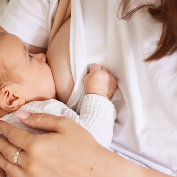 Imagem focada em um bebê no seio da mãe, alimentado-se com leite materno. Ele tem a pele clara e o cabelo castanho claro. Está com um body branco e o olhar em direção a mãe, que veste uma camiseta branca.