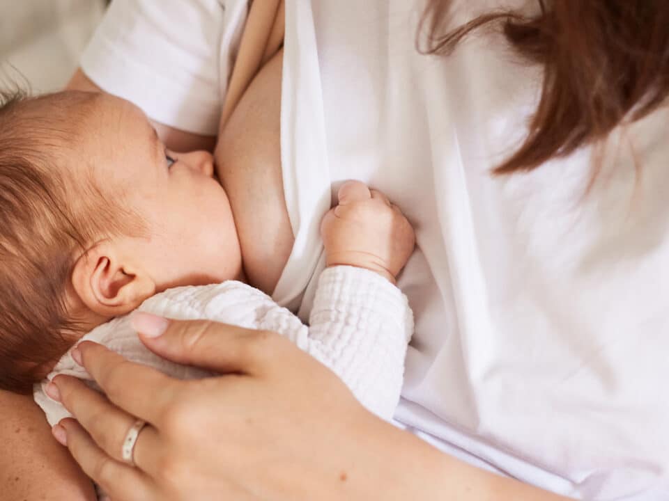 Imagem focada em um bebê no seio da mãe, alimentado-se com leite materno. Ele tem a pele clara e o cabelo castanho claro. Está com um body branco e o olhar em direção a mãe, que veste uma camiseta branca.