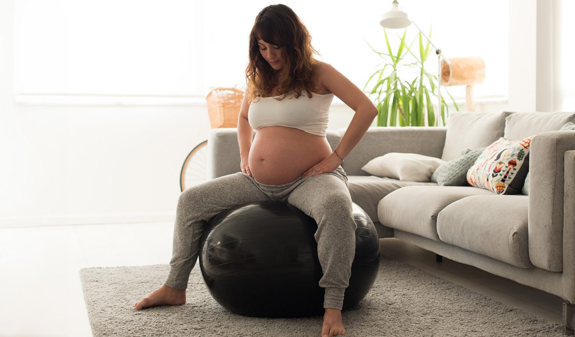 Imagem de uma gestante sentada em uma bola de pilates. Ela está com as mãos nos quadris e parece estar realizando algum exercício para aliviar o desconforto de uma das fases do trabalho de parto. Ela está em uma sala de estar. Veste calça de moletom cinza claro e um top branco.