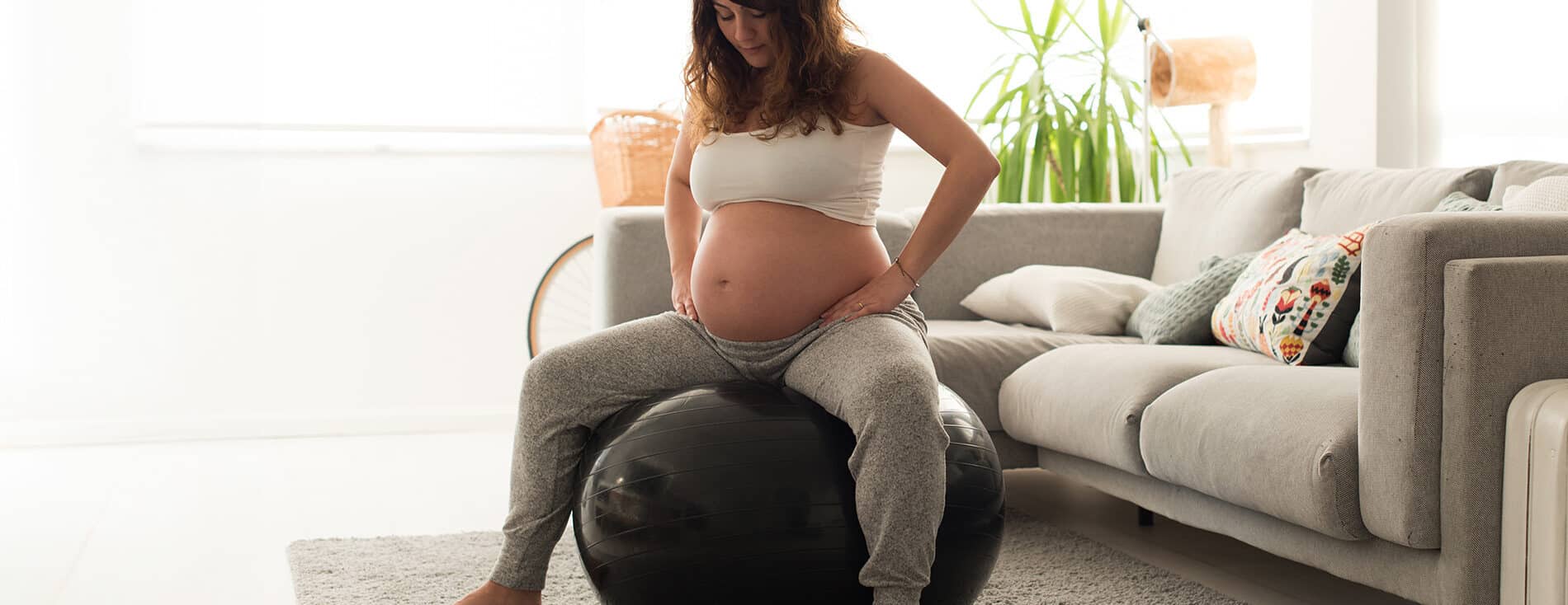 Imagem de uma gestante sentada em uma bola de pilates. Ela está com as mãos nos quadris e parece estar realizando algum exercício para aliviar o desconforto de uma das fases do trabalho de parto. Ela está em uma sala de estar. Veste calça de moletom cinza claro e um top branco.