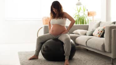 Imagem de uma gestante sentada em uma bola de pilates. Ela está com as mãos nos quadris e parece estar realizando algum exercício para aliviar o desconforto de uma das fases do trabalho de parto. Ela está em uma sala de estar. Veste calça de moletom cinza claro e um top branco.