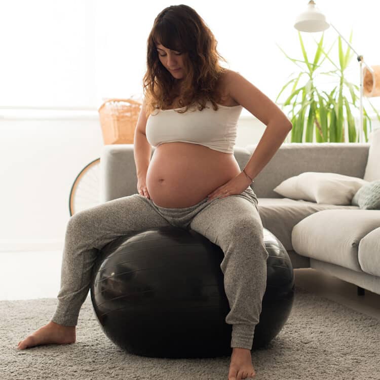 Imagem de uma gestante sentada em uma bola de pilates. Ela está com as mãos nos quadris e parece estar realizando algum exercício para aliviar o desconforto de uma das fases do trabalho de parto. Ela está em uma sala de estar. Veste calça de moletom cinza claro e um top branco.