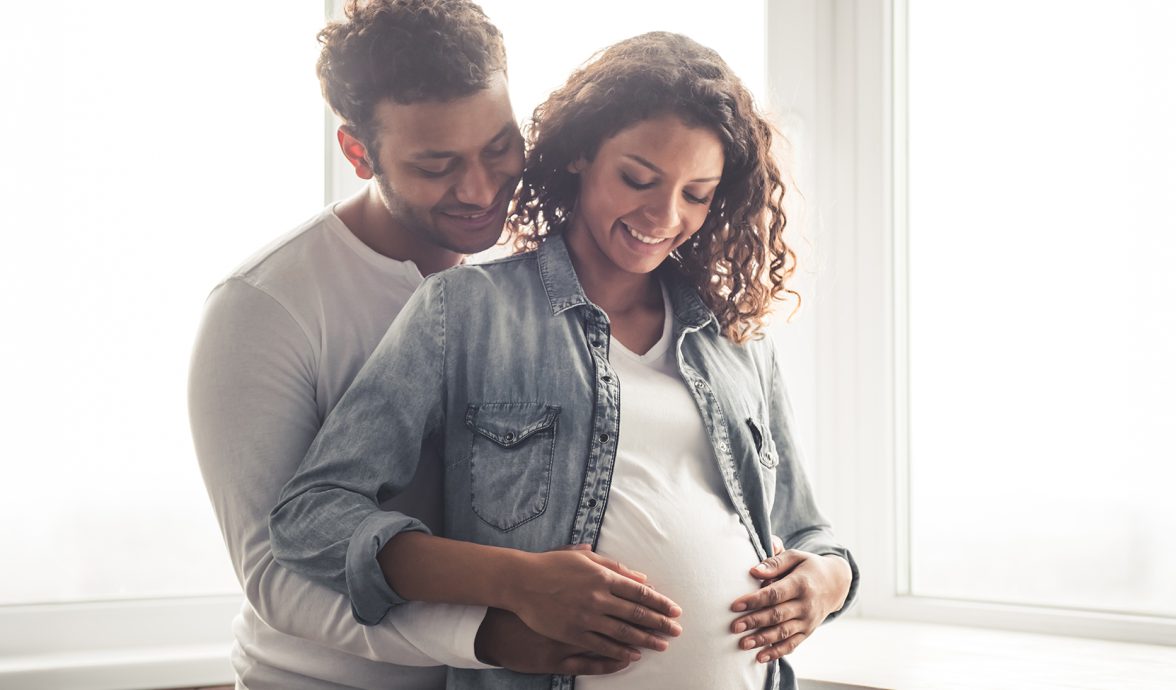 Imagem de um casal. amos tem a pele negra e estão sorrindo. Ele está abraçando a mulher e ambos estão com as mãos na barriga dela, que está grávida, demonstrando a fertilidade do casal.
