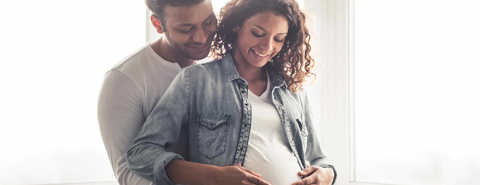 Imagem de um casal. amos tem a pele negra e estão sorrindo. Ele está abraçando a mulher e ambos estão com as mãos na barriga dela, que está grávida, demonstrando a fertilidade do casal.