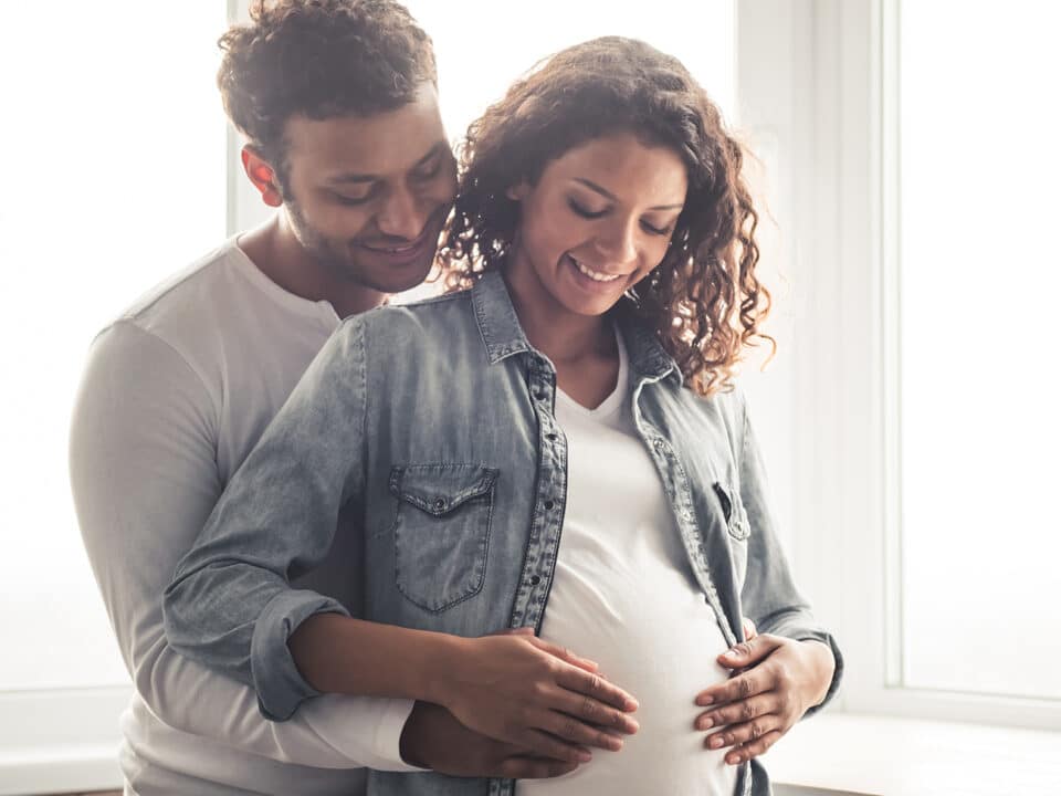 Imagem de um casal. amos tem a pele negra e estão sorrindo. Ele está abraçando a mulher e ambos estão com as mãos na barriga dela, que está grávida, demonstrando a fertilidade do casal.