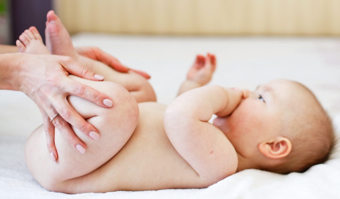Foto de um bebê com aproximadamente 3 meses. Ele tem a pele clara, pouco cabelo e está deitado sobre uma cama de lonçol branco. As mãos da mãe estão dobrando as perninhas do bebê, um movimento para aliviar a disquesia do lactente.