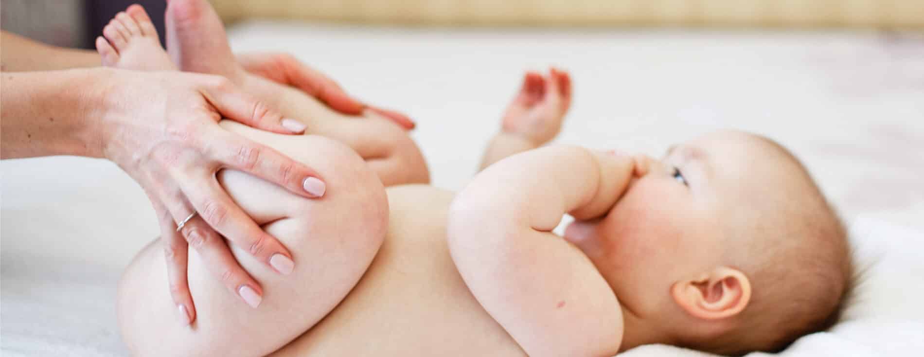 Foto de um bebê com aproximadamente 3 meses. Ele tem a pele clara, pouco cabelo e está deitado sobre uma cama de lonçol branco. As mãos da mãe estão dobrando as perninhas do bebê, um movimento para aliviar a disquesia do lactente.