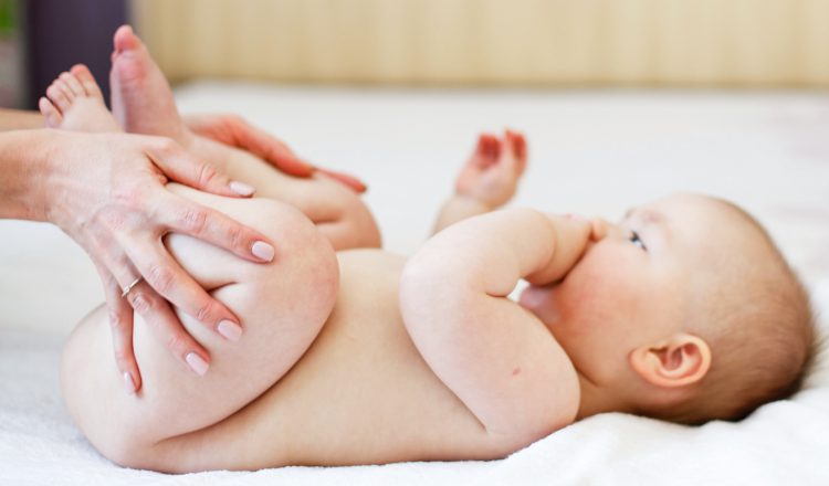 Foto de um bebê com aproximadamente 3 meses. Ele tem a pele clara, pouco cabelo e está deitado sobre uma cama de lonçol branco. As mãos da mãe estão dobrando as perninhas do bebê, um movimento para aliviar a disquesia do lactente.