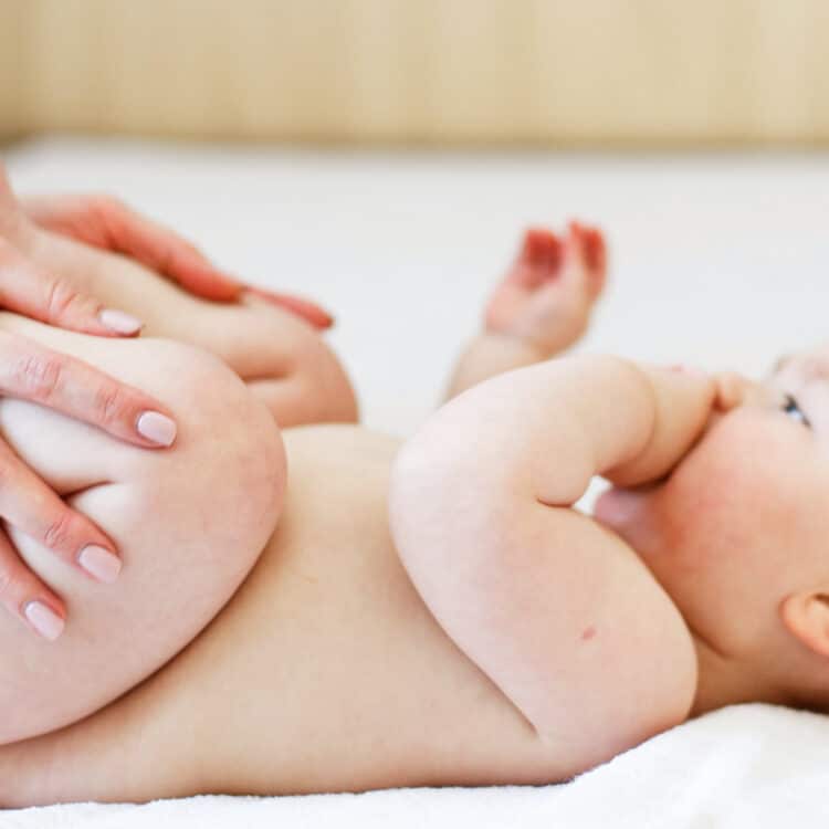 Foto de um bebê com aproximadamente 3 meses. Ele tem a pele clara, pouco cabelo e está deitado sobre uma cama de lonçol branco. As mãos da mãe estão dobrando as perninhas do bebê, um movimento para aliviar a disquesia do lactente.