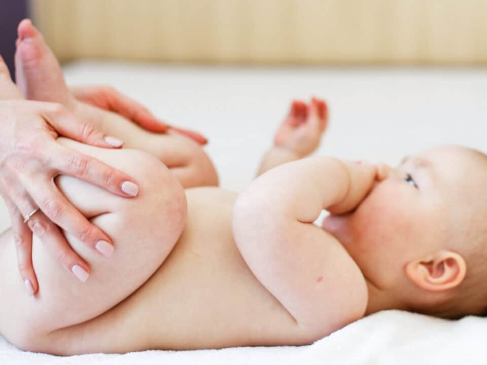 Foto de um bebê com aproximadamente 3 meses. Ele tem a pele clara, pouco cabelo e está deitado sobre uma cama de lonçol branco. As mãos da mãe estão dobrando as perninhas do bebê, um movimento para aliviar a disquesia do lactente.