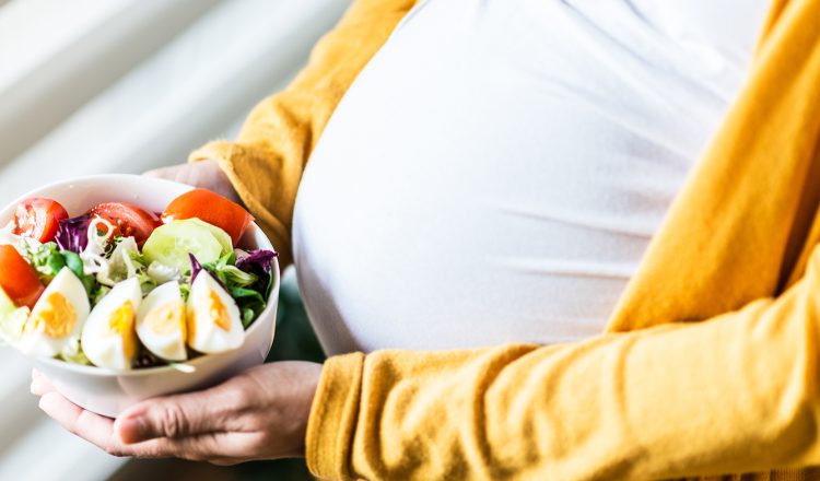 Imagem fechada mostrando a barriga e as mãos de uma gestante. Entre as mãos está um bowl com salada e ovos, uma importante fonte de proteína na gravidez.