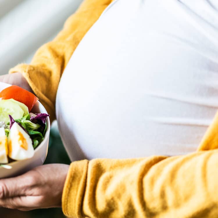 Imagem fechada mostrando a barriga e as mãos de uma gestante. Entre as mãos está um bowl com salada e ovos, uma importante fonte de proteína na gravidez.