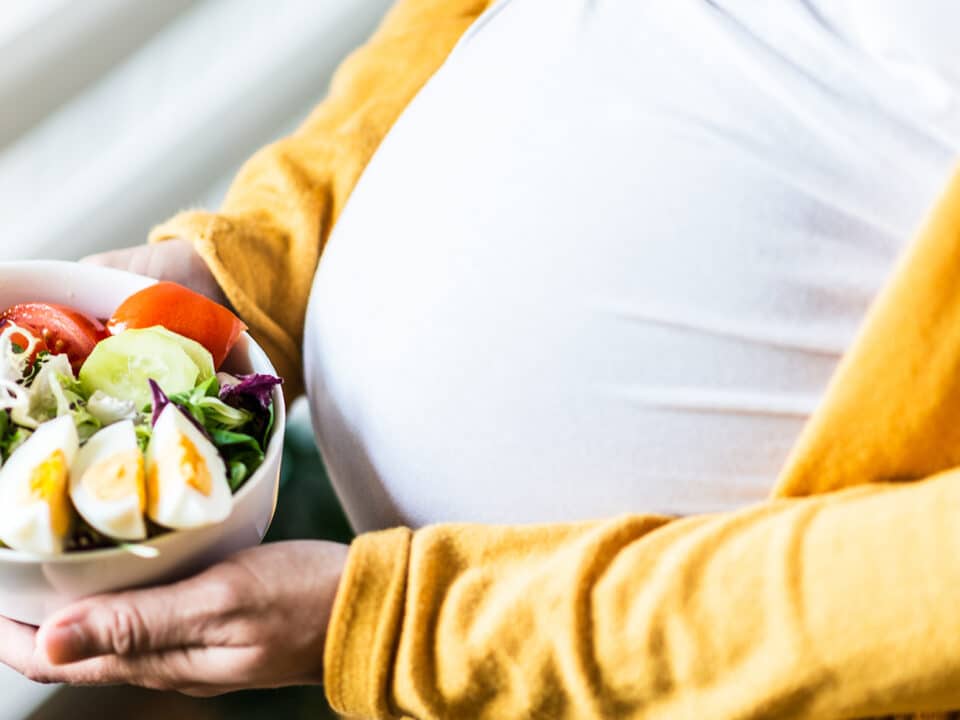 Imagem fechada mostrando a barriga e as mãos de uma gestante. Entre as mãos está um bowl com salada e ovos, uma importante fonte de proteína na gravidez.