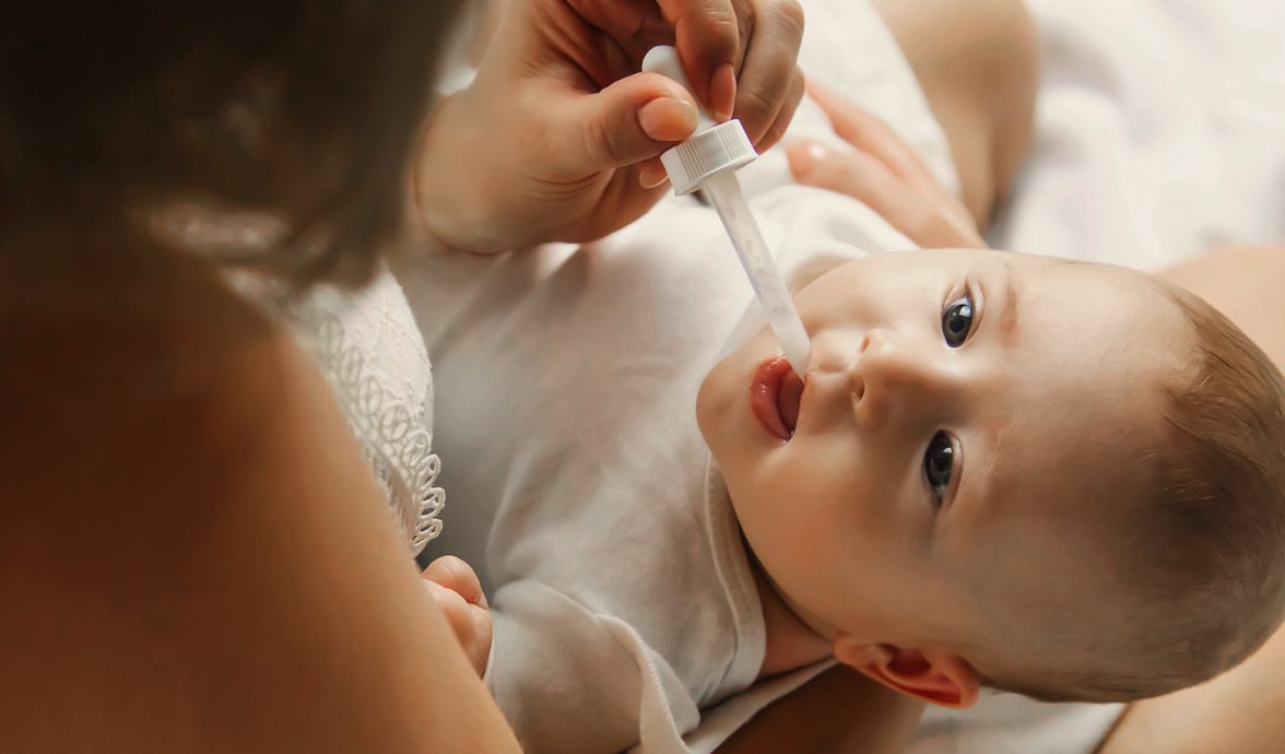 Imagem de um bebê deitado no colo da mãe. Ela está suplementando vitamina D para o bebê por meio de um aplicador conta-gotas.