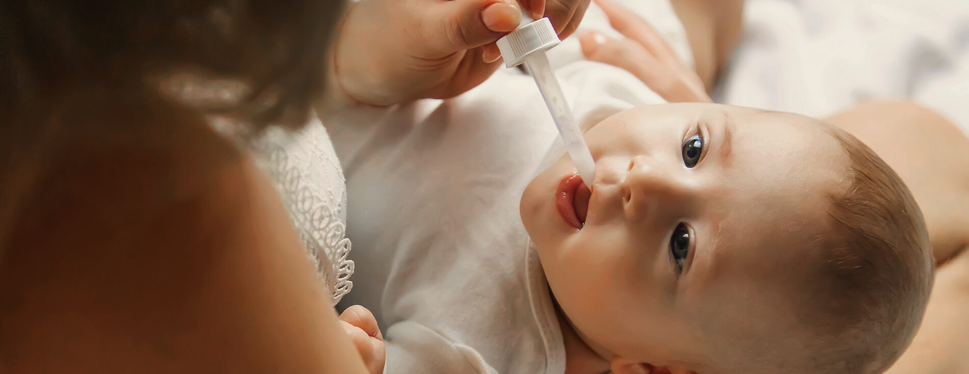 Imagem de um bebê deitado no colo da mãe. Ela está suplementando vitamina D para o bebê por meio de um aplicador conta-gotas.