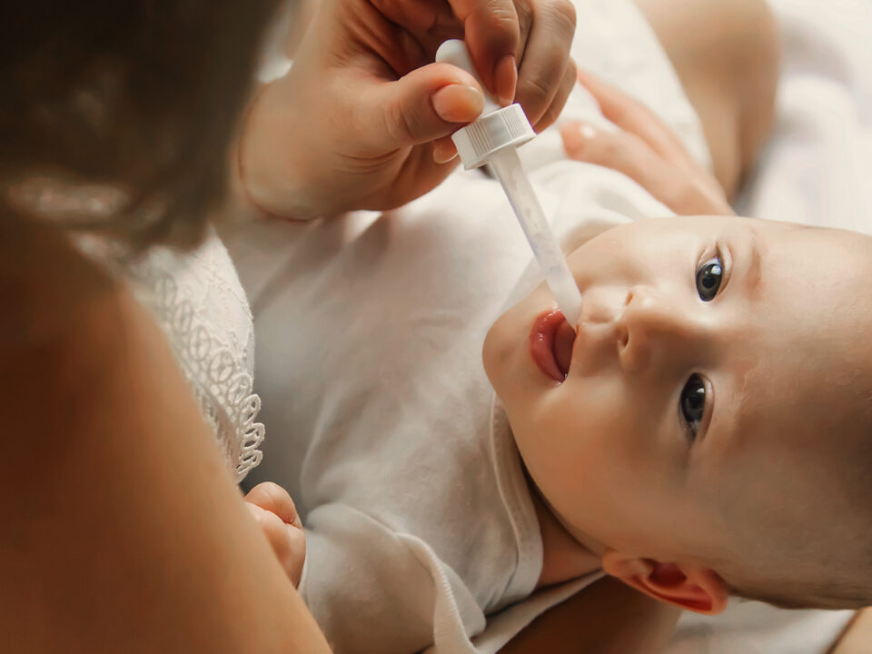 Imagem de um bebê deitado no colo da mãe. Ela está suplementando vitamina D para o bebê por meio de um aplicador conta-gotas.