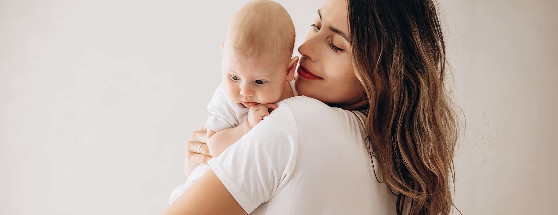 Imagem de uma mãe com seu bebê no colo. Ela está de costas e o bebê está apoiado em um dos seus ombros. Ela olha sorridente para o pequeno.