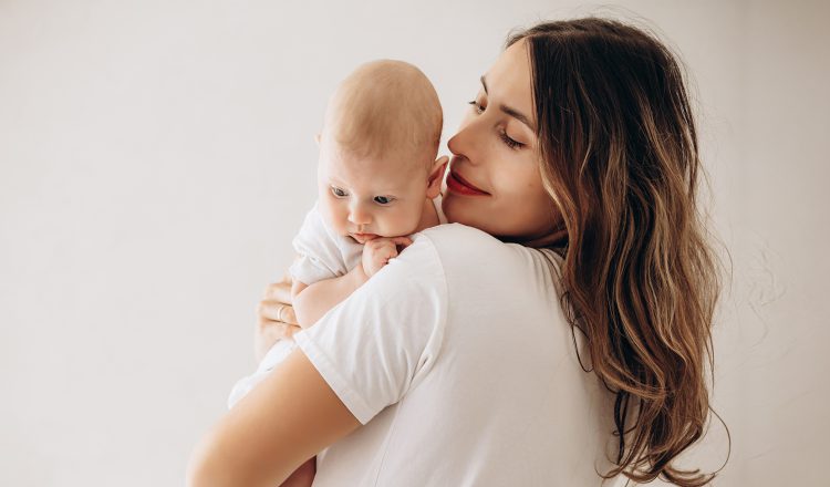 Imagem de uma mãe com seu bebê no colo. Ela está de costas e o bebê está apoiado em um dos seus ombros. Ela olha sorridente para o pequeno.