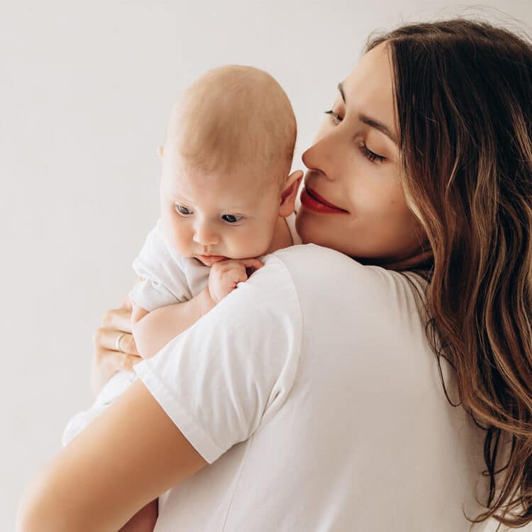 Imagem de uma mãe com seu bebê no colo. Ela está de costas e o bebê está apoiado em um dos seus ombros. Ela olha sorridente para o pequeno.