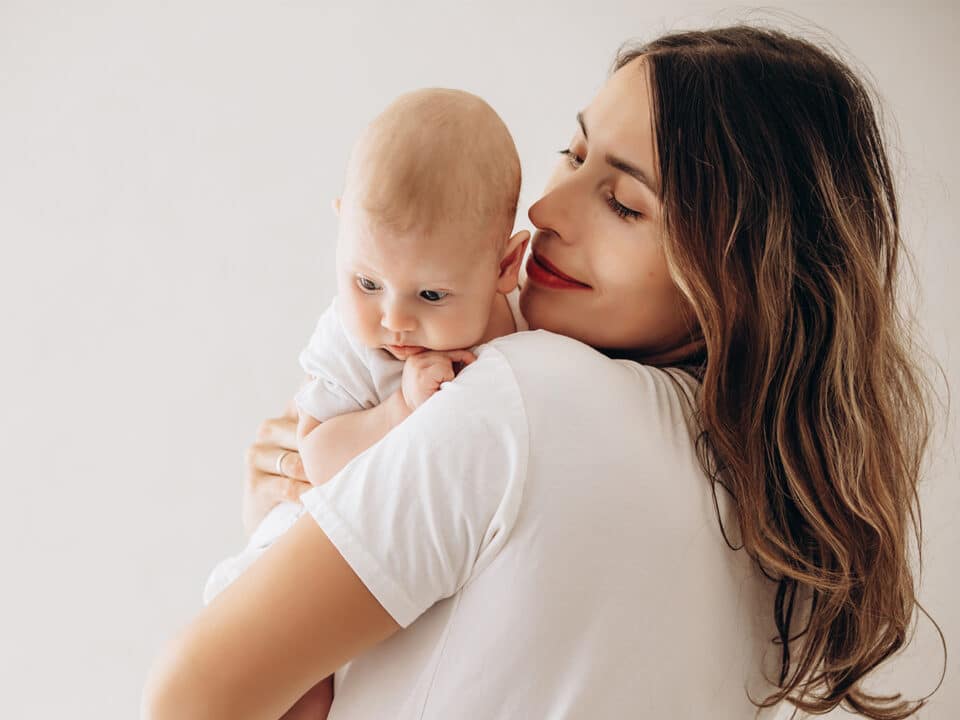 Imagem de uma mãe com seu bebê no colo. Ela está de costas e o bebê está apoiado em um dos seus ombros. Ela olha sorridente para o pequeno.