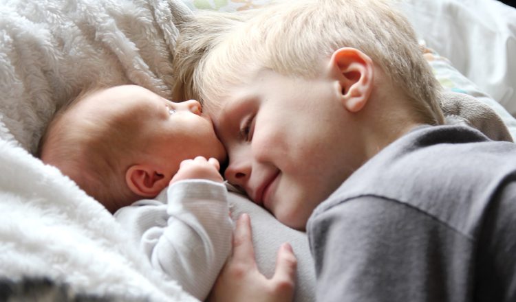 Imagem de um bebê e uma criança. Ambos tem a pele e o cabelo claros e estão deitados. O irmão mais velho está abraçando o bebê.
