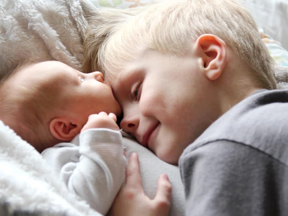 Imagem de um bebê e uma criança. Ambos tem a pele e o cabelo claros e estão deitados. O irmão mais velho está abraçando o bebê.