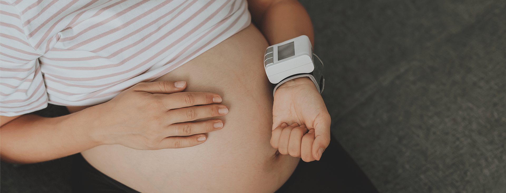 Imagem fechada na barriga de uma gestante. Ela está com uma das mãos sobre a barriga e a outra aferindo a pressão arterial, um indicador importante relacionado a pré-eclâmpsia.
