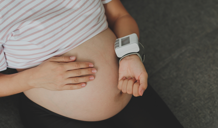 Imagem fechada na barriga de uma gestante. Ela está com uma das mãos sobre a barriga e a outra aferindo a pressão arterial, um indicador importante relacionado a pré-eclâmpsia.