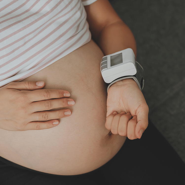 Imagem fechada na barriga de uma gestante. Ela está com uma das mãos sobre a barriga e a outra aferindo a pressão arterial, um indicador importante relacionado a pré-eclâmpsia.