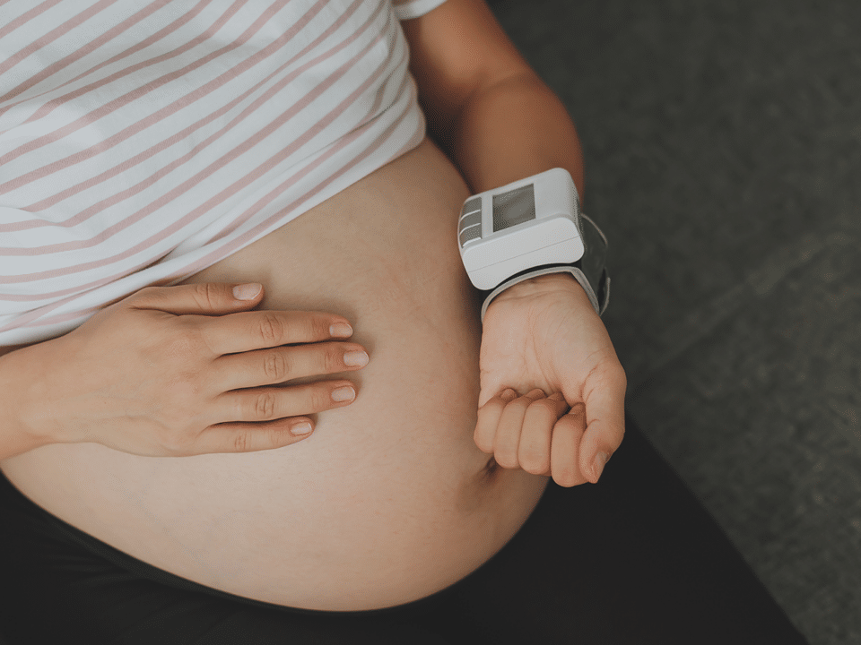Imagem fechada na barriga de uma gestante. Ela está com uma das mãos sobre a barriga e a outra aferindo a pressão arterial, um indicador importante relacionado a pré-eclâmpsia.
