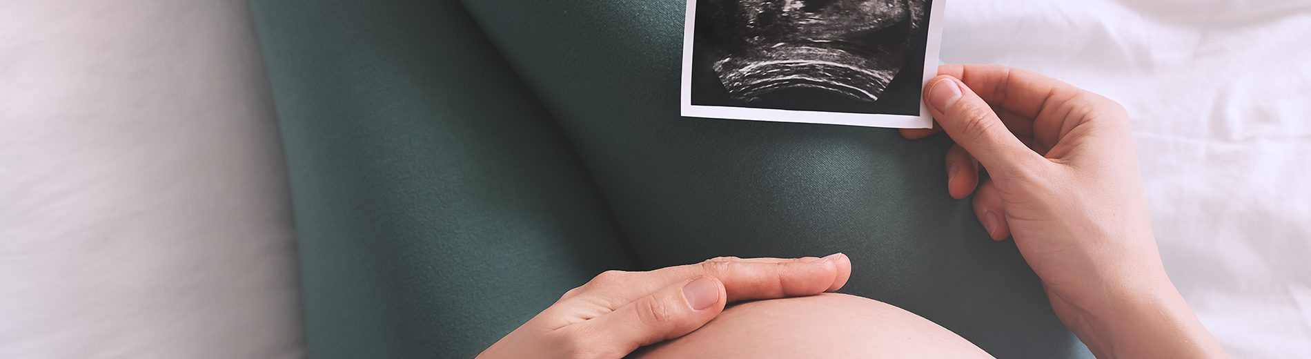 Imagem fechada da barriga de uma gestante. Ela está sentada, com uma das mãos na barriga e a outra segurando uma imagem de ultrassom, exame realizado para identificar o percentil fetal do bebê.