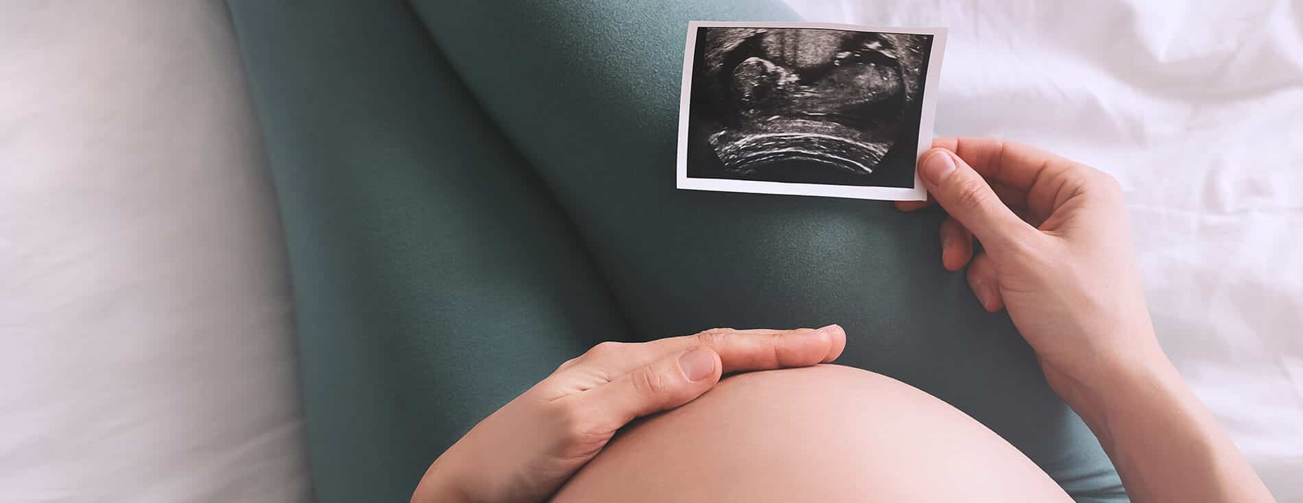 Imagem fechada da barriga de uma gestante. Ela está sentada, com uma das mãos na barriga e a outra segurando uma imagem de ultrassom, exame realizado para identificar o percentil fetal do bebê.