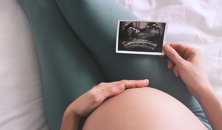 Imagem fechada da barriga de uma gestante. Ela está sentada, com uma das mãos na barriga e a outra segurando uma imagem de ultrassom, exame realizado para identificar o percentil fetal do bebê.