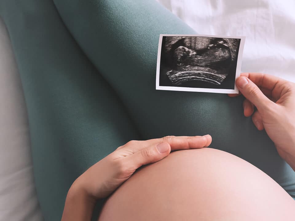 Imagem fechada da barriga de uma gestante. Ela está sentada, com uma das mãos na barriga e a outra segurando uma imagem de ultrassom, exame realizado para identificar o percentil fetal do bebê.