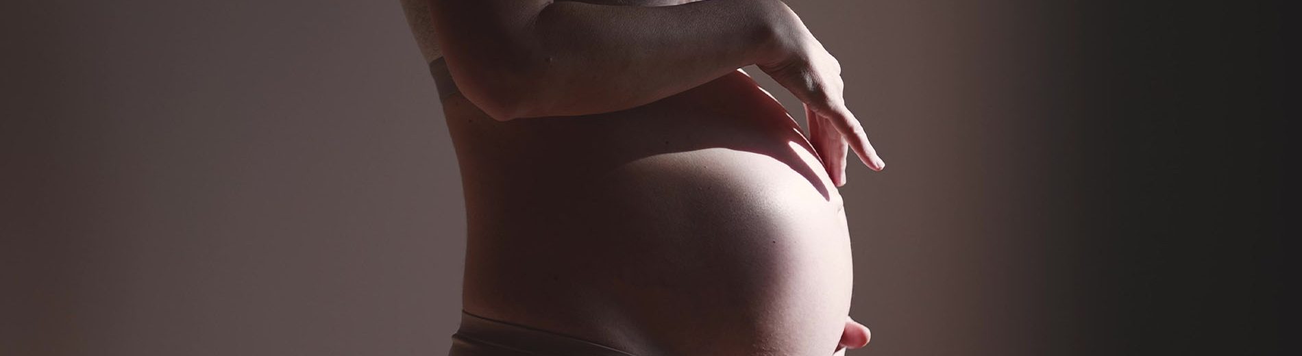 Imagem do corpo de uma gestante. Ela está de perfil e as suas mãos envolvem a barriga. A imagem está escura com um feixe de luz incidindo sobre a barriga.