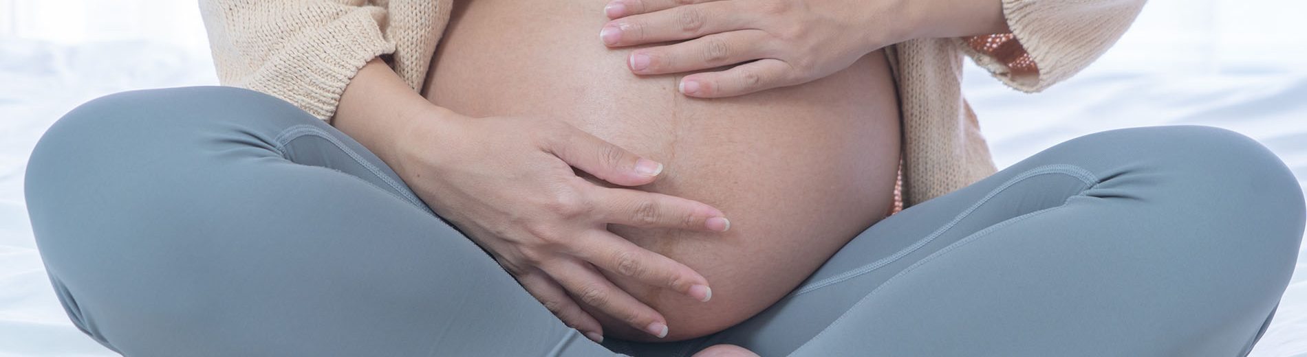 Imagem do corpo de uma gestante. Ela tem a pele clara, veste um top branco, uma caça de fginástica cinza e um cardigan creme. Está com as pernas em posição de borboleta e acariciando a barriga com as mãos.