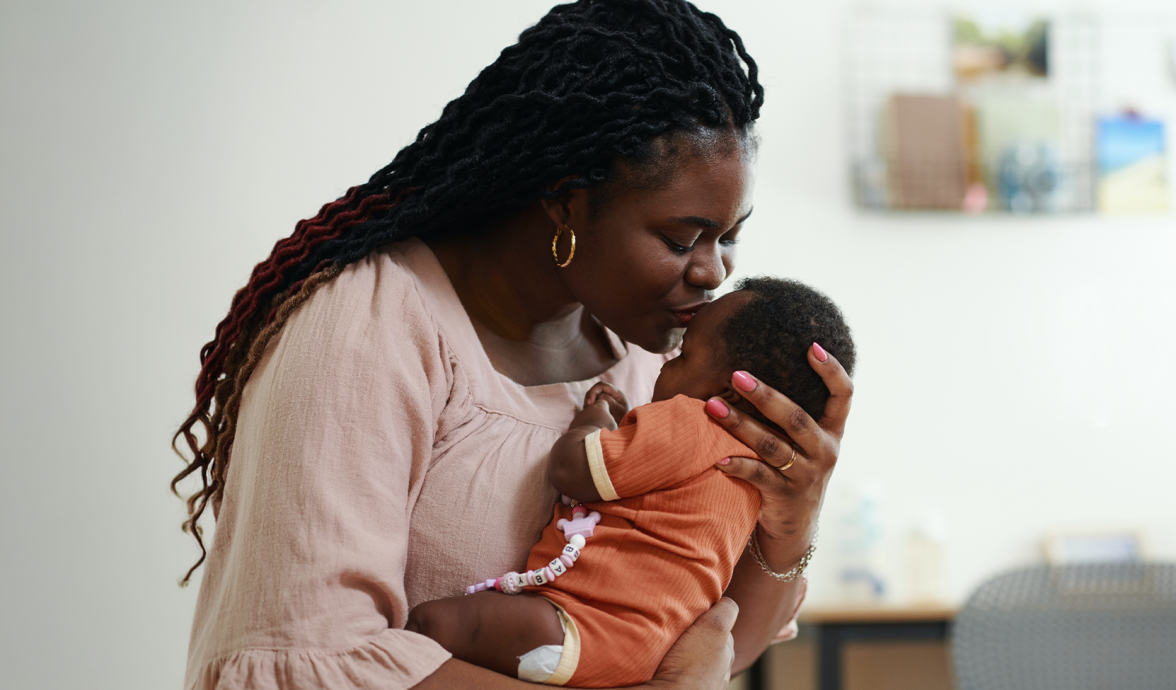 Imagem de uma mulher dando colo para o bebê. Ela tem a pele negra, os cabelos escuros, encaracolados e longos. Está de olhos fechados e dando um beijo na testa do bebê, que também tem a pele negra e veste um body laranja.